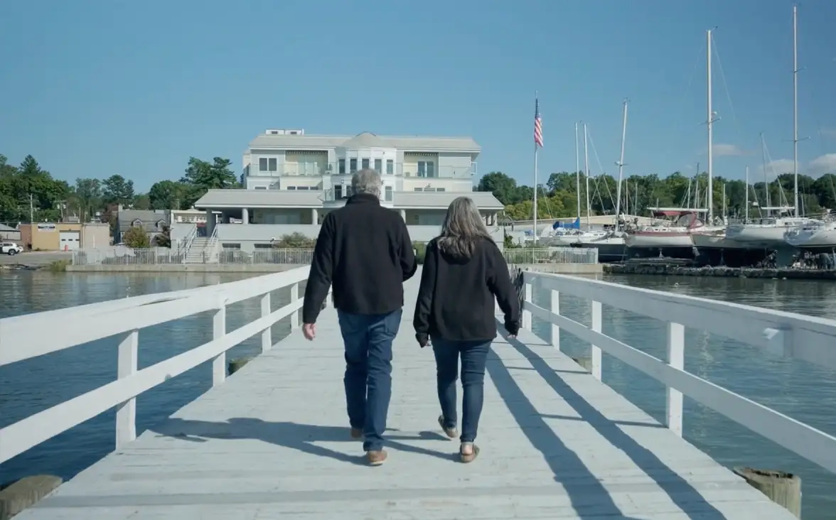 Two people walking down a white pier towards a building and boats on a sunny waterfront.