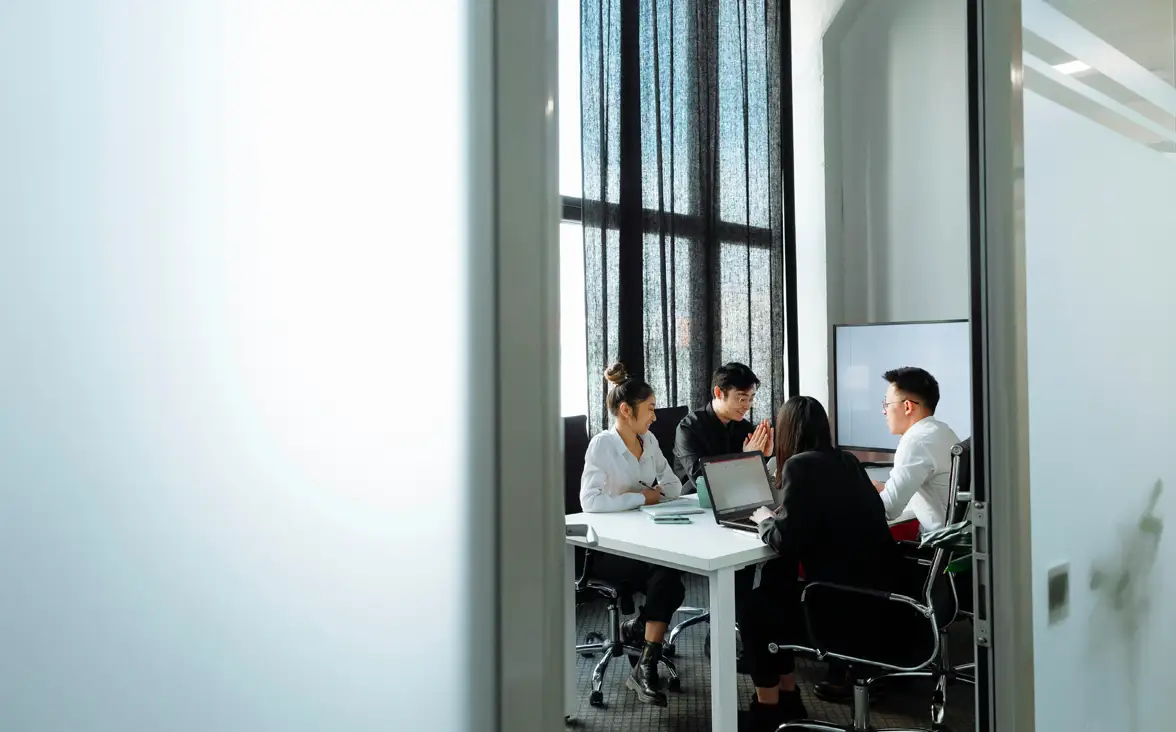 Four people engaged in a meeting around a table with laptops in a brightly lit conference room.