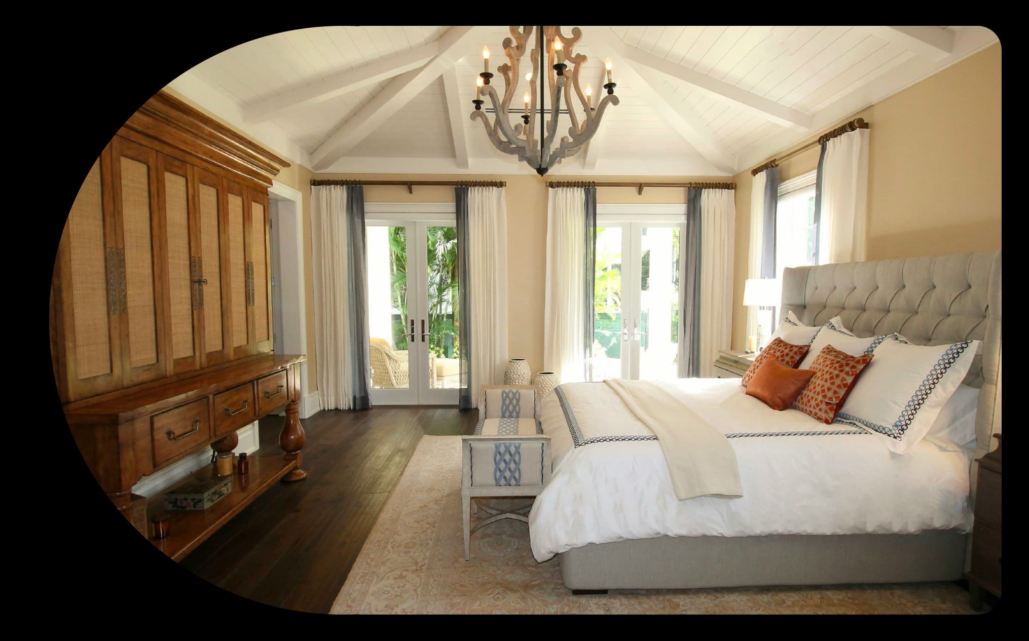 Spacious bedroom with vaulted ceiling, chandelier, upholstered bed, wooden armoire, and bright windows.