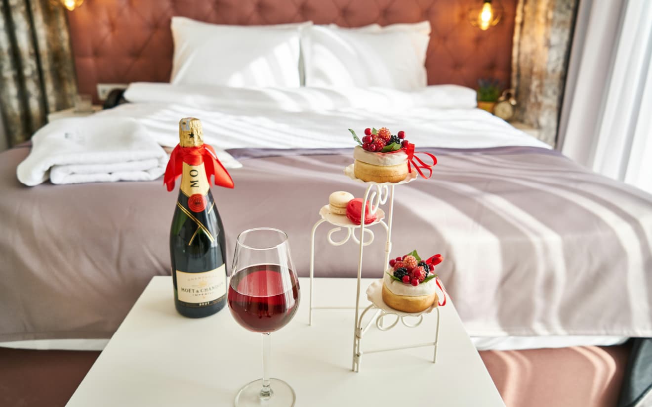 Hotel room bed with wine bottle, filled glass, and pastries arranged decoratively on a small table.