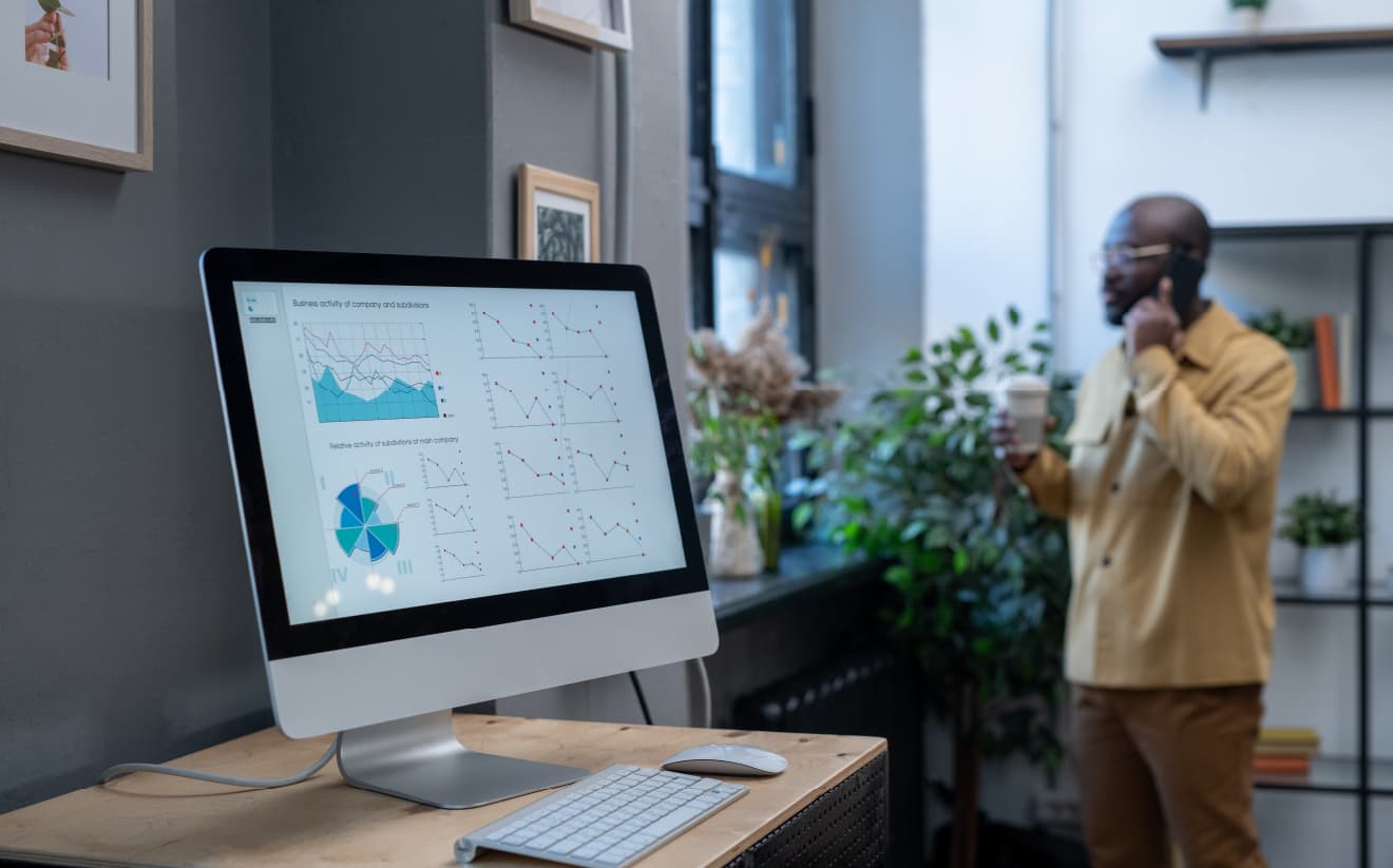 Computer showing business charts; man in background holding coffee and on phone in modern office.