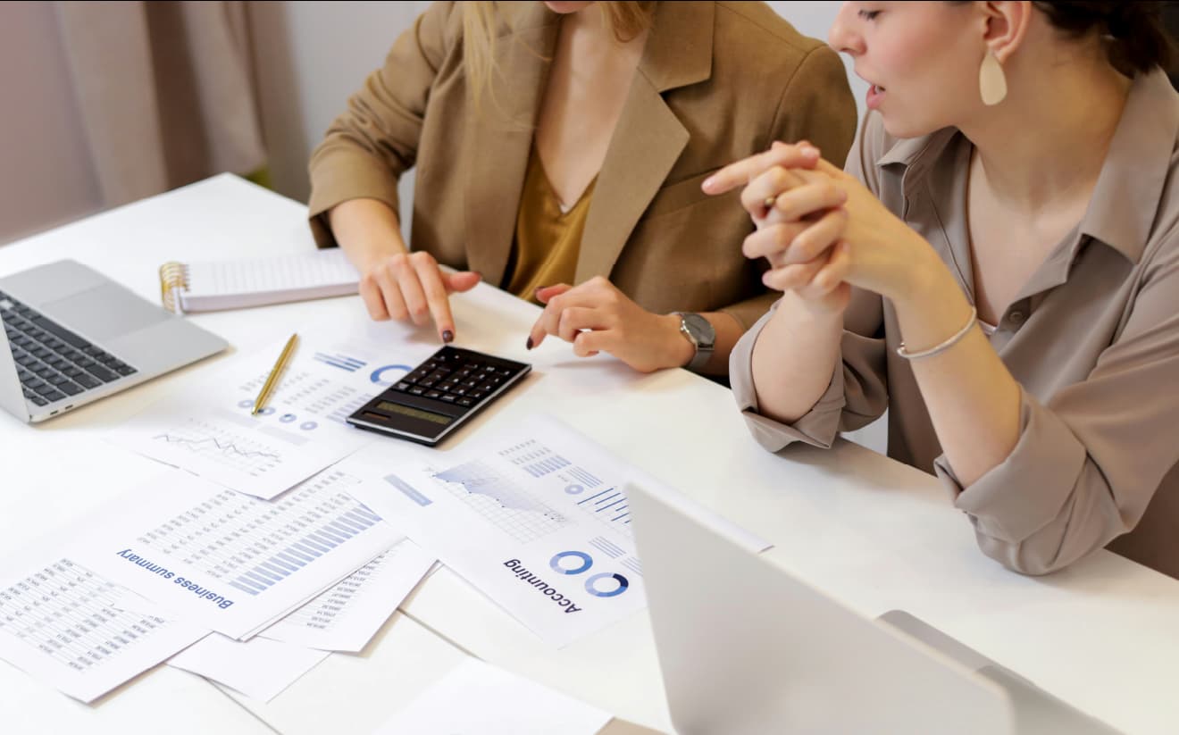 Two people discussing and reviewing financial documents, using a calculator at a desk with laptops and charts.