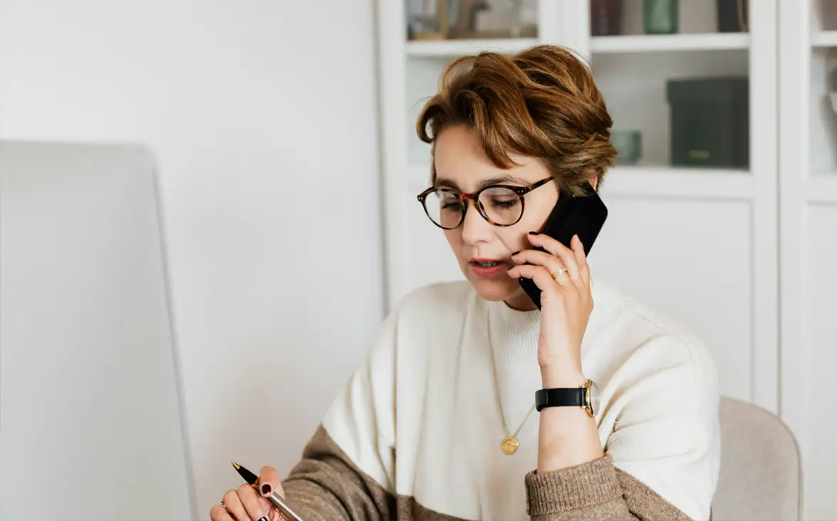 Woman wearing glasses talks on the phone and takes notes in an office setting.