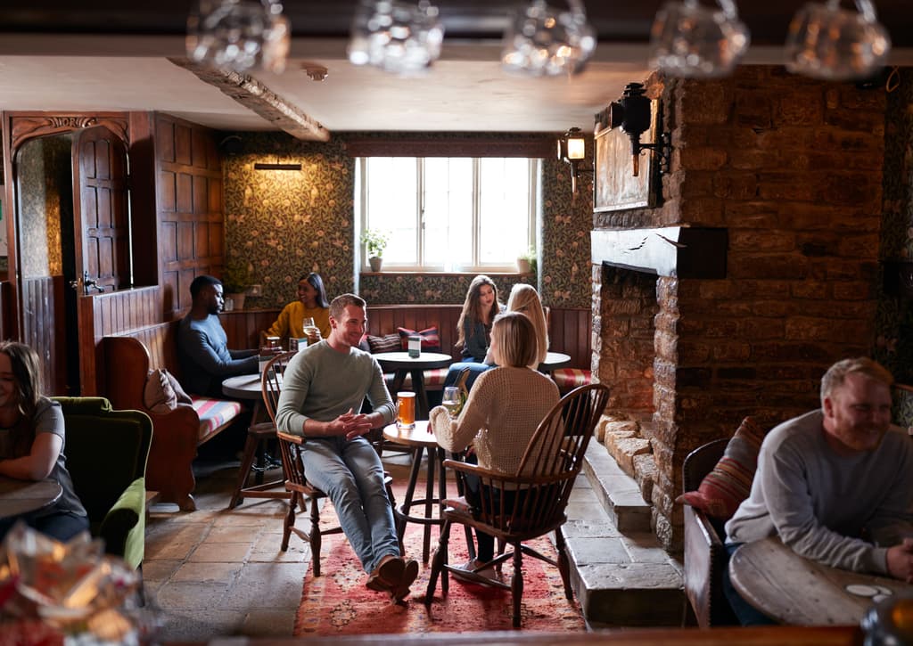 A lively interior scene of a traditional, cozy inn, featuring several small groups of diverse people sitting at tables, chatting and enjoying drinks, with a couple smiling in the foreground.