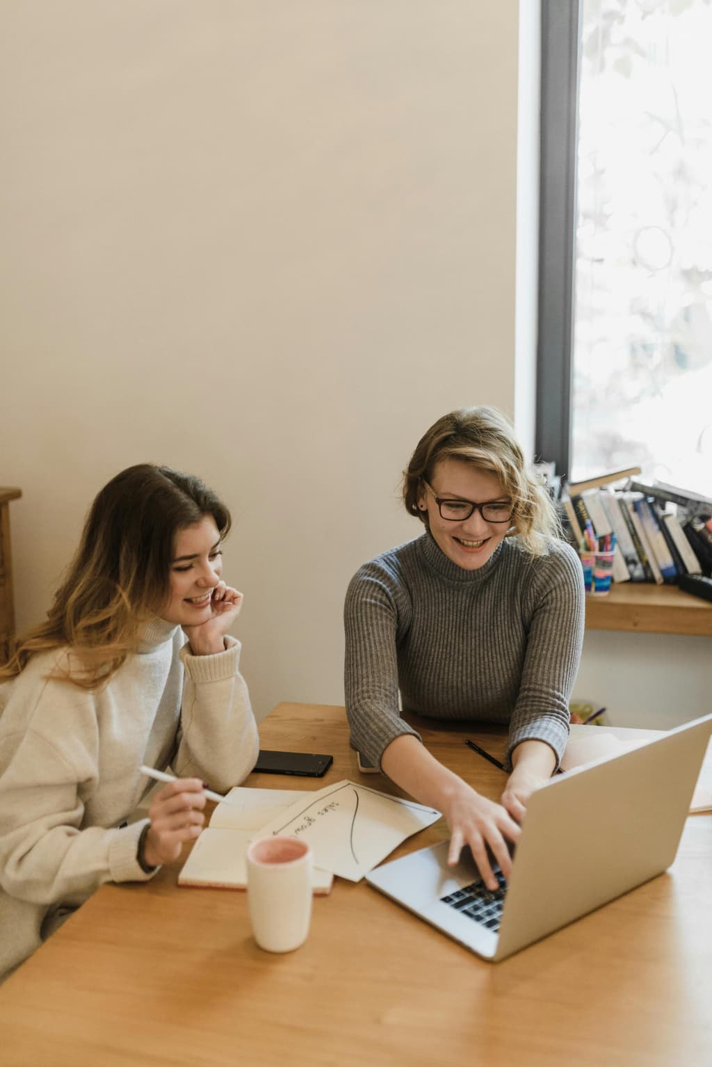 Two young women smiling and collaborating on a laptop and notebook at a wooden table in a brightly lit room.