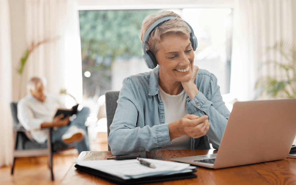 A female experienced lodging professional attends a virtual conference with her laptop and headphones. A man in the background reads.