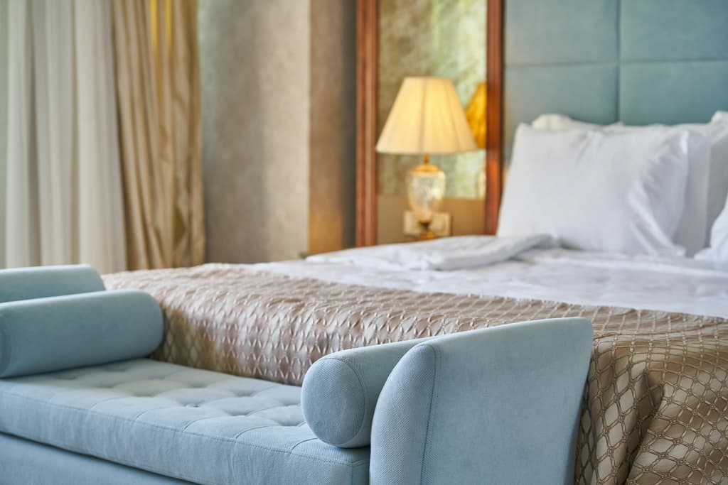 A hotel-style bedroom featuring a light blue velvet upholstered bench at the foot of a bed with a tufted teal headboard.
