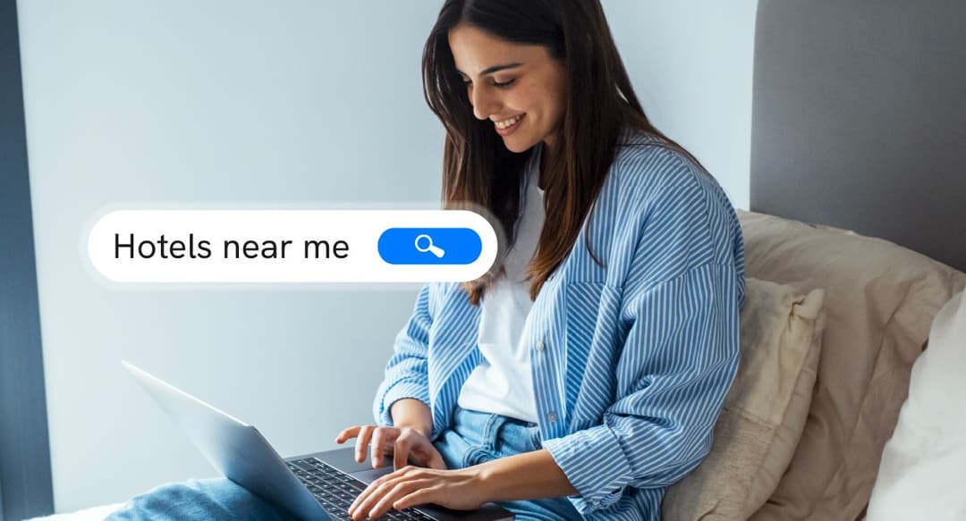A smiling woman with a laptop, featuring a superimposed search bar that reads "Hotels near me.