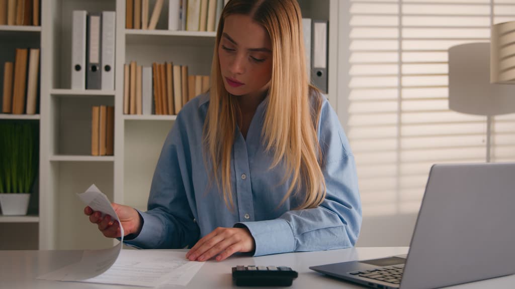 A focused blonde woman in a light blue shirt reviews papers and uses a calculator at a white desk with a laptop, in front of a bookshelf and window blinds.